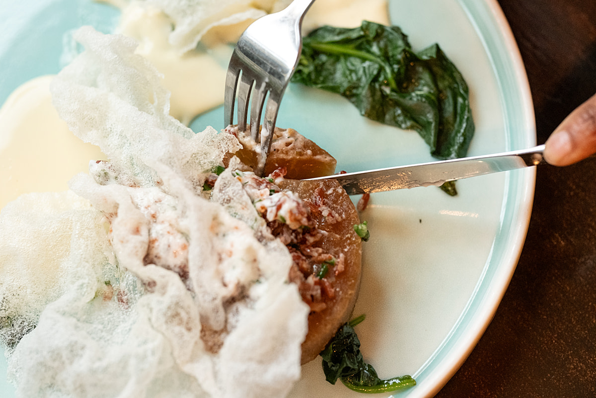 A fork and knife cut into a vegan seafood specialty item at Studio Gauthier. The dish consists of of kohlrabi, rice and a samphire kaffir seasoned vegan butter topped off with a rice paper flower chip. Studio Gauthier, London, England, UK, 2024. Nathalie Merle / We Animals