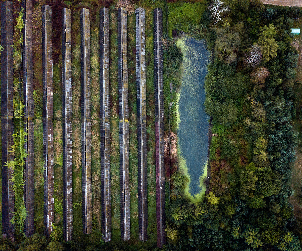 An aerial view of a shuttered mink farm, closed for several years and located deep in a forest near a town. The facility once housed tens of thousands of mink who were raised for their fur. Vegetation grows over what remains of the partially dismantled structures. Poland, 2019. Andrew Skowron / We Animals