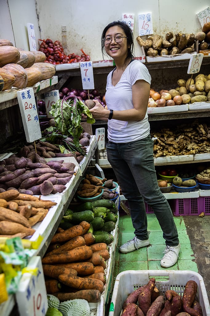 Jah Ying Chung, a food researcher in China, smiles as she selects vegetables from the display inside a store that sells produce. Jah Ying is the co-founder of The Good Growth Co., which researches Chinese consumers' attitudes toward food and works in the plant-based/alternative protein and animal welfare spaces. Hong Kong, China, 2022. #unboundproject / We Animals Media